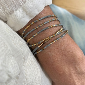 Op de afbeelding: Een stapel grijze kralenarmbanden met een gouden accent. De armbanden zijn op elkaar gelegd op de pols van een persoon.