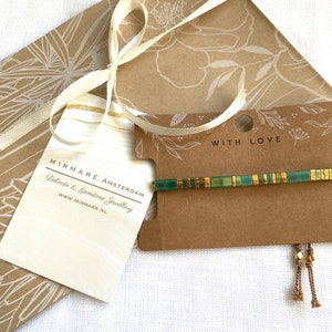 Puede incluir: Un brazalete de cuentas verdes y doradas con un dije de borla. El brazalete est&aacute; empaquetado en una tarjeta con el texto "Mirmare Amsterdam, Joyer&iacute;a Delicada y Piedras Preciosas, www.mirmare.nl" y "Con amor".