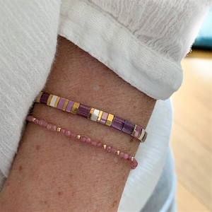 May include: Two bracelets worn together. One bracelet features a repeating pattern of rectangular purple, white, and metallic gold beads. The second bracelet consists of small, faceted pink gemstone beads interspersed with tiny, round gold-tone metal beads.