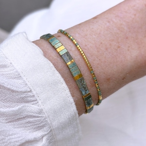 Könnte beinhalten: Zwei Armbänder, die zusammen getragen werden. Ein Armband besteht aus breiteren, rechteckigen, marmorierten blaugrünen und goldfarbenen Perlen. Das zweite, dünnere Armband besteht aus kleinen, abwechselnden zylindrischen blaugrünen und goldfarbenen Perlen.
