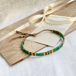 Puede incluir: Un brazalete de piedras preciosas verde y dorado con un cord&oacute;n marr&oacute;n y cuentas doradas. El brazalete est&aacute; sobre una bolsa de papel marr&oacute;n con una etiqueta blanca que dice "Mirmare Amsterdam, Joyer&iacute;a delicada y de piedras preciosas".