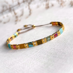 May include: A delicate bracelet featuring alternating rectangular beads in turquoise, iridescent pink, and brown, separated by small gold-toned metallic spacers. The piece includes a thin adjustable cord closure with gold-toned end beads.