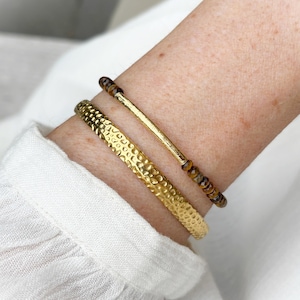 Può includere: Due braccialetti color oro. Uno è un polsino largo e piatto con una superficie martellata e testurizzata. Il secondo è una catenina sottile con una barra centrale dorata affiancata da piccole perle rotonde in pietra occhio di tigre marrone.