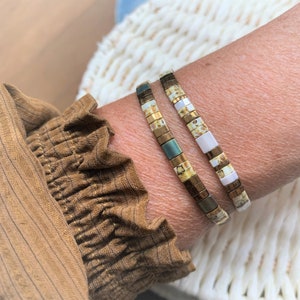 Op de afbeelding: Twee elastische armbanden bestaande uit kleine, rechthoekige, platte kralen. De kralen hebben een mix van metallic brons, matgroen en glanzend witte afwerkingen, gerangschikt in afwisselende patronen langs het elastische koord van elke armband.