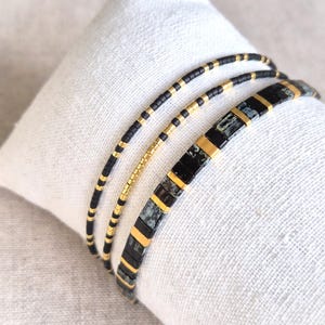 May include: A set of three bracelets featuring thin, cylindrical beads in alternating black and gold colors. The third bracelet is wider, composed of rectangular black, gold, and mottled gray stone-like beads arranged in a repeating pattern.