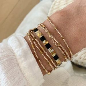 Könnte beinhalten: Ein Set aus goldenen und schwarzen Perlenarmbändern mit einem gemusterten Design. Die Armbänder sind auf dem Handgelenk einer Person gestapelt.