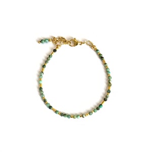 Könnte beinhalten: Ein zartes Armband mit kleinen, facettierten, grün-braun gesprenkelten Perlen, die sich mit dünnen goldfarbenen zylindrischen Zwischenstücken abwechseln. Das Schmuckstück hat einen goldfarbenen Karabinerverschluss und eine kurze Verlängerungskette.