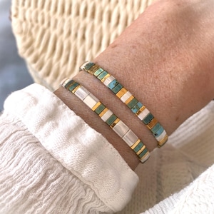 Pode incluir: Duas pulseiras sobrepostas com contas retangulares em padrões alternados de ouro metálico, branco mate e azul turquesa. As contas estão dispostas numa sequência repetitiva, criando um design linear e estruturado à volta do pulso.