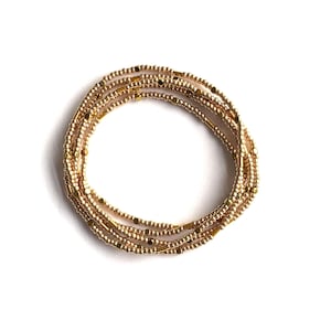 May include: Gold beaded body jewelry. The jewelry is made of small, round beads and rectangular beads strung together. The beads are a shiny gold colour. The jewelry is arranged in a circular shape on a white background.
