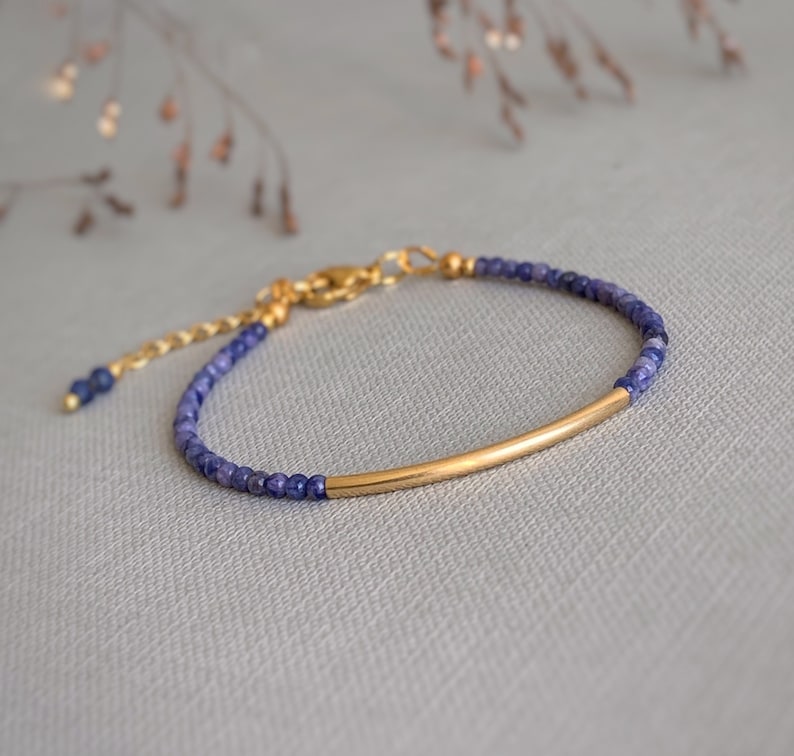 Puede incluir: Una pulsera delicada con peque&ntilde;as cuentas facetadas de color azul oscuro y una pieza central de tubo de metal dorado curvado. La pulsera incluye una cadena extensora de metal dorado con un cierre de mosquet&oacute;n y una cuenta azul al final.
