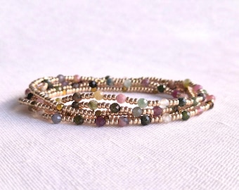 May include: A multi-strand bracelet composed of small metallic gold-toned beads interspersed with tiny, faceted, multi-colored gemstone beads in shades of pink, green, brown, and clear, arranged in a circular, layered coil design.