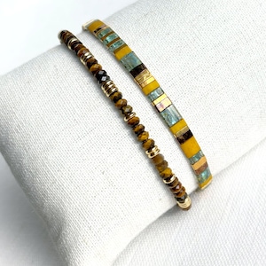 Könnte beinhalten: Ein Set aus zwei Armbändern. Ein Armband besteht aus kleinen, facettierten braunen Tigeraugenperlen, die mit goldfarbenen Metallzwischenstücken kombiniert sind. Das zweite Armband zeigt flache, rechteckige Perlen in Senfgelb und Petrol.