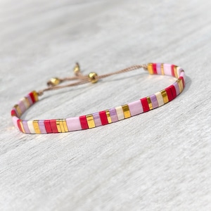 May include: A bracelet featuring a repeating pattern of flat, rectangular beads in shades of red, light pink, lavender, and metallic gold. The bracelet includes a thin brown cord with two small gold-toned beads at the adjustable closure mechanism.
