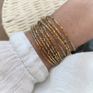 May include: A multi-strand bracelet with small, speckled beige and gold beads. The bracelet is wrapped around a wrist, highlighting the delicate design and colour contrast. The gold beads add a touch of elegance to the accessory.