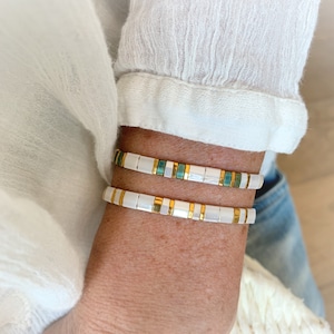 Op de afbeelding: Twee witte tegelarmbanden met gouden en groene accenten. De armbanden zijn gestapeld op de pols van een persoon.