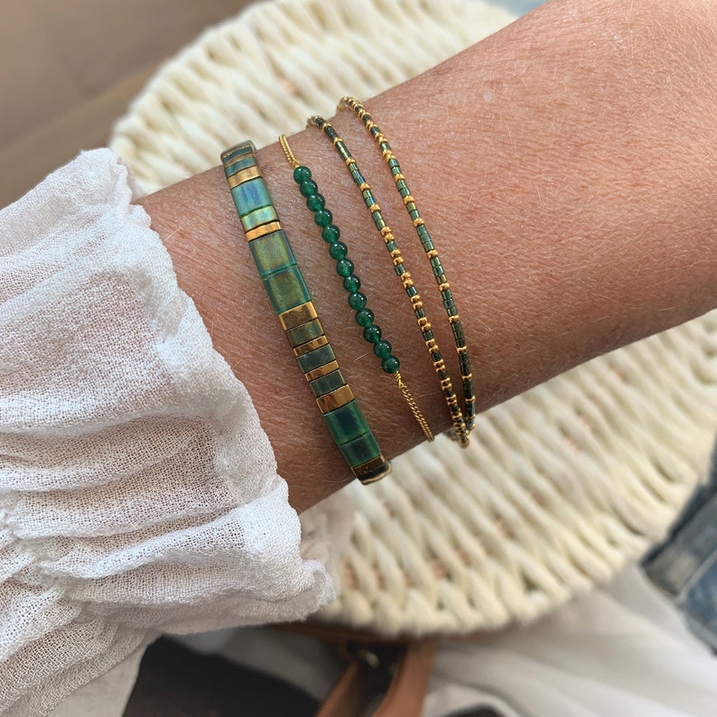 Puede incluir: Un conjunto de cuatro pulseras con cuentas verdes y doradas. Las pulseras est&aacute;n apiladas en la mu&ntilde;eca de una persona.