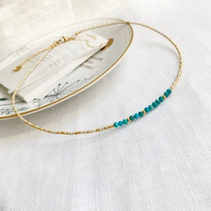 May include: A delicate necklace featuring a strand of tiny cream-colored seed beads interspersed with small gold-toned metallic beads. A section of the necklace showcases a series of faceted turquoise-colored beads accented by gold spacers.