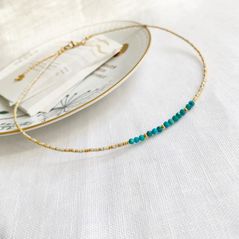 May include: A delicate necklace featuring a strand of tiny cream-colored seed beads interspersed with small gold-toned metallic beads. A section of the necklace showcases a series of faceted turquoise-colored beads accented by gold spacers.