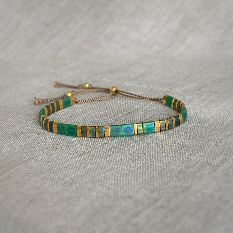 Puede incluir: Un brazalete de cuentas verdes y doradas con cierre de cord&oacute;n marr&oacute;n. El brazalete est&aacute; hecho de peque&ntilde;as cuentas cuadradas con un sutil brillo iridiscente.