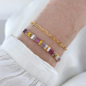 May include: Two bracelets on a wrist. One is a gold chain link bracelet. The other is a beaded bracelet with purple, white, and gold square beads. The bracelets are on a light skin tone wrist, with a white shirt in the background.