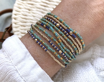 Puede incluir: Una pila de múltiples pulseras de cuentas con un patrón repetitivo de pequeñas cuentas de color turquesa, azul oscuro y oro metálico. Las pulseras están dispuestas en capas, mostrando una mezcla de cuentas redondas lisas y detalles dorados.