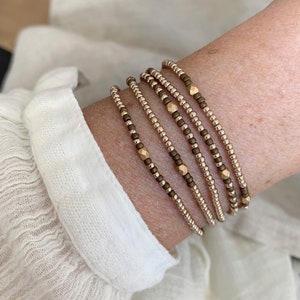 May include: A set of six delicate bracelets featuring a mix of gold and brown beads. The bracelets are stacked on a person's wrist, creating a layered look.