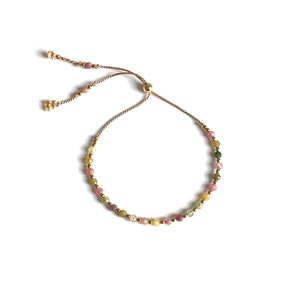 Peut inclure: Un bracelet réglable délicat composé d'un cordon fin marron avec un fermoir coulissant à perle dorée. Le bracelet est orné de petites perles en pierre gemme facettées multicolores dans des tons rose, jaune et vert, alternées avec de minuscules perles intercalaires métalliques dorées.