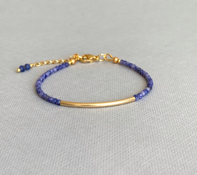Puede incluir: Una pulsera delicada con peque&ntilde;as cuentas facetadas de color azul oscuro ensartadas en un cord&oacute;n fino. El dise&ntilde;o incluye un tubo de metal dorado, liso y curvado, centrado entre las cuentas, con un cierre de mosquet&oacute;n dorado y una cadena ajustable.