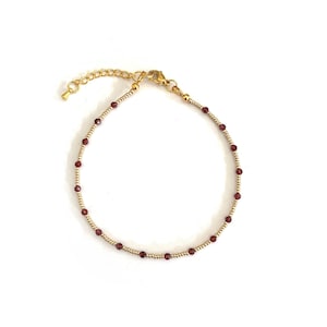 Op de afbeelding: Een verfijnde armband met een herhalend patroon van kleine, ronde, donkerrode granaatkralen afgewisseld met piepkleine, gepolijste goudkleurige metalen kralen. De armband is voorzien van een goudkleurige karabijnsluiting en een verstelbare ketting.