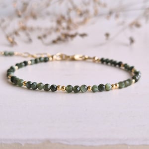 Könnte beinhalten: Ein zartes Armband mit kleinen, facettierten dunkelgrünen Edelsteinperlen, die mit winzigen, polierten goldfarbenen Metallkugeln durchsetzt sind. Das Armband verfügt über eine goldfarbene Metallkettenverlängerung und einen Karabinerverschluss.