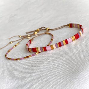 May include: Two beaded bracelets with adjustable closures. The larger bracelet features red, pink, gold, and lavender rectangular beads. The smaller bracelet has a similar design with smaller beads. Both bracelets are on a white surface.
