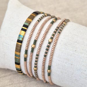 Op de afbeelding: Een set van vijf armbanden met verschillende soorten kralen. Eén armband bestaat uit platte, rechthoekige tegeltjes in zwart, goud en blauwgroen. De andere vier armbanden bestaan uit kleine, ronde, metallic roségouden en matte olijfgroene kralen.