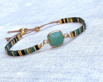 Puede incluir: Una pulsera con una piedra central cuadrada de color verde claro engastada en metal dorado, flanqueada por segmentos alternos de cuentas de color verde azulado, negro y dorado. La banda está hecha de un cordón marrón fino.