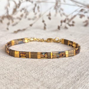 Op de afbeelding: Een armband met een herhalend patroon van metallic gouden, mat bronskleurige en iriserende amberkleurige rechthoekige kralen. Het sieraad is afgewerkt met een goudkleurige metalen karabijnsluiting en een kort stukje bijpassende goudkleurige ketting.