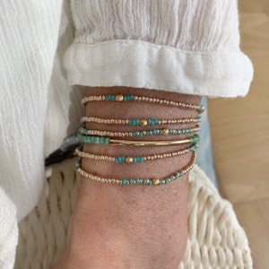Könnte beinhalten: Ein Set aus sechs gestapelten Armbändern mit kleinen metallisch goldfarbenen Perlen und türkisfarbenen Perlen. Ein Armband enthält einen glatten, horizontalen goldfarbenen Metallstab, während andere gelegentlich facettierte goldfarbene Akzentperlen aufweisen.