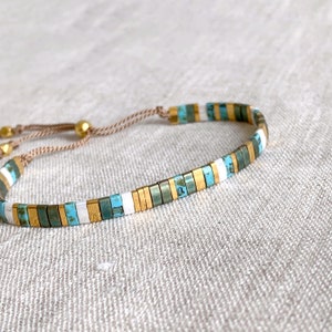 May include: A delicate bracelet featuring a repeating pattern of small, rectangular beads in turquoise, metallic gold, and white. The beads are strung on a thin, light brown cord with adjustable gold-toned metal beads at the end of the pull strings.