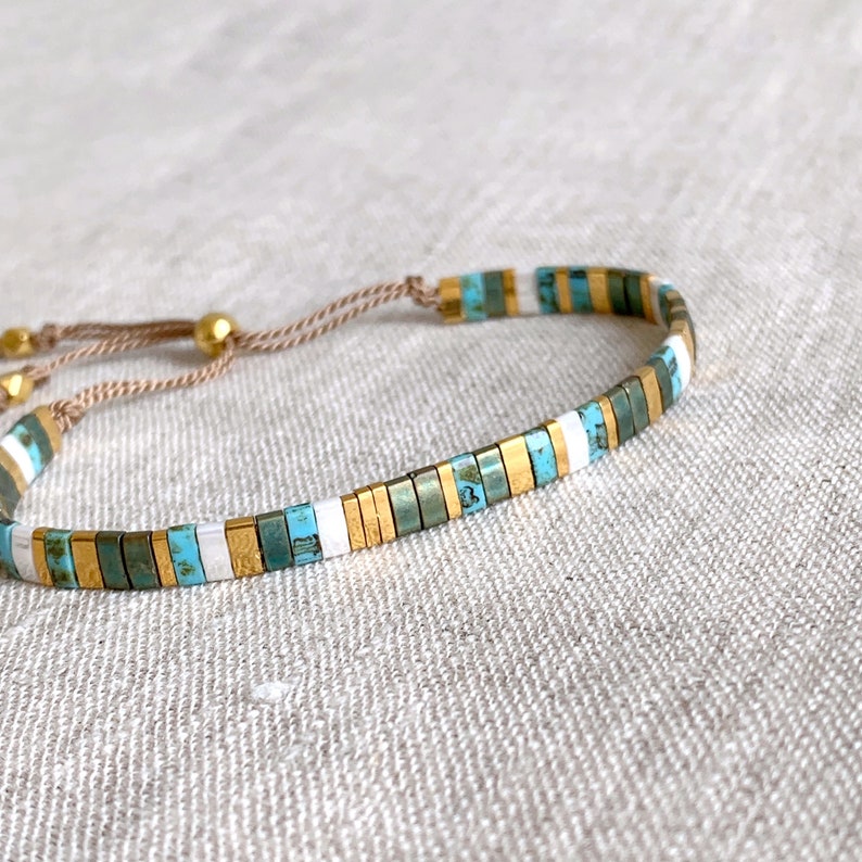 May include: A delicate bracelet featuring a repeating pattern of small, rectangular beads in turquoise, metallic gold, and white. The beads are strung on a thin, light brown cord with adjustable gold-toned metal beads at the end of the pull strings.