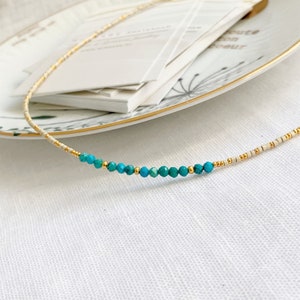 May include: A delicate beaded necklace featuring a sequence of small, cylindrical cream-colored beads interspersed with tiny metallic gold spacers and a central row of faceted, round turquoise-colored gemstone beads with gold accents.