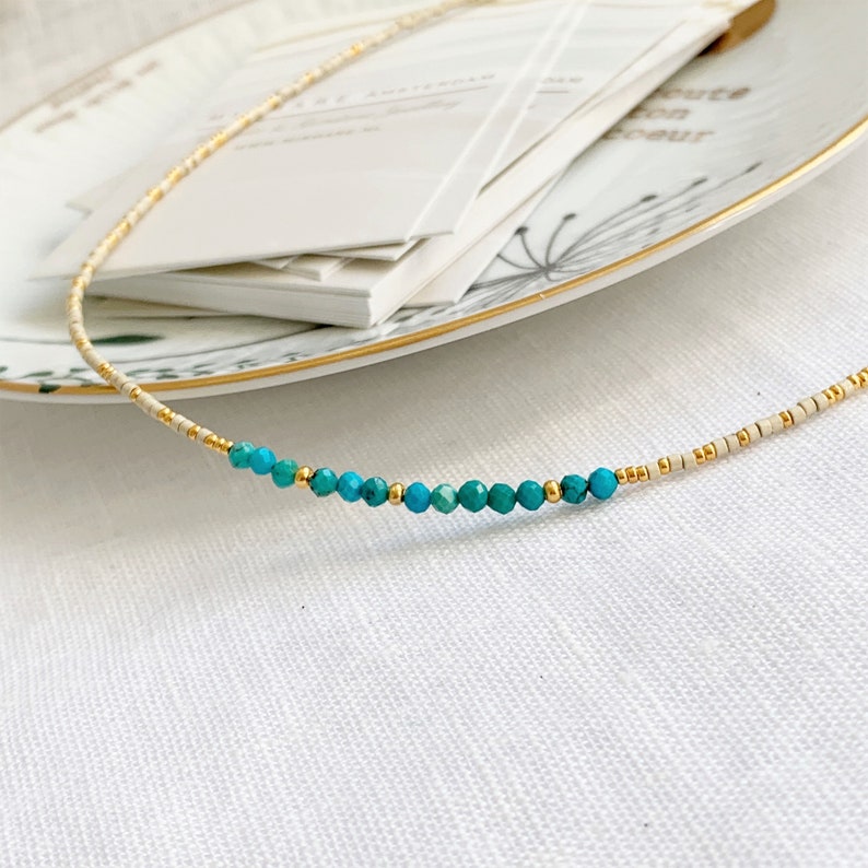 May include: A delicate beaded necklace featuring a sequence of small, cylindrical cream-colored beads interspersed with tiny metallic gold spacers and a central row of faceted, round turquoise-colored gemstone beads with gold accents.