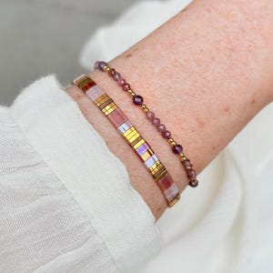 Può includere: Due braccialetti indossati insieme. Uno è un braccialetto a piastrelle rettangolari piatte con segmenti alternati in oro metallizzato, rosa iridescente e rosa traslucido. Il secondo è un delicato braccialetto di perline con piccole pietre viola sfaccettate e perline dorate.