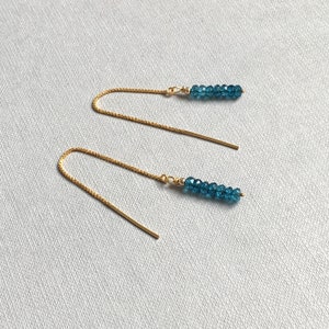 Könnte beinhalten: Ein Paar goldfarbene Ohrringe zum Einfädeln. Jeder Ohrring hat eine Kette, die sich in einen geraden Stab erstreckt, mit einer Ansammlung kleiner, facettierter, teal-farbener Perlen am Ende. Die Ohrringe werden auf einer hellgrauen Oberfläche präsentiert.