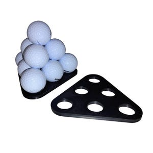 May include: A black triangular golf ball holder with six holes, next to a stack of nine white golf balls. The golf balls are arranged in a pyramid shape on a matching black base.