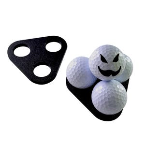 May include: Black golf ball holder with three golf balls. One golf ball has a black face design. The holder is black and has three holes. The golf balls are white.