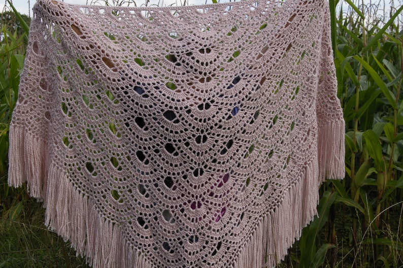 May include: A delicate, crocheted shawl in a soft pink color with a lacy pattern and fringe trim. The shawl is draped over a person's arms and is photographed against a backdrop of green foliage.