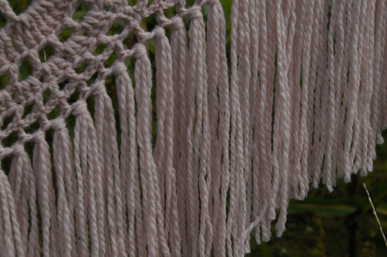May include: A crocheted shawl with a delicate pattern and long, flowing fringe. The shawl is made of a soft, light pink yarn.