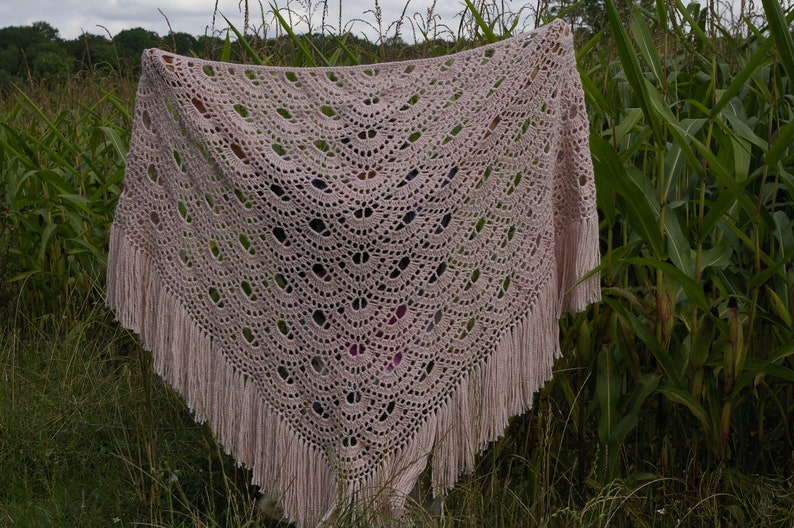 May include: A delicate, crocheted shawl in a soft pink color with a fringe border. The shawl is made with a lacy pattern and is displayed against a backdrop of green foliage.