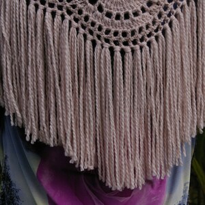 May include: A crocheted shawl with a circular design and a fringe border. The shawl is made of a light pink yarn.