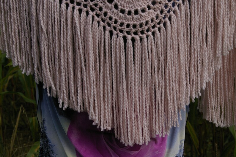 May include: A crocheted shawl with a circular design and a fringe border. The shawl is made of a light pink yarn.