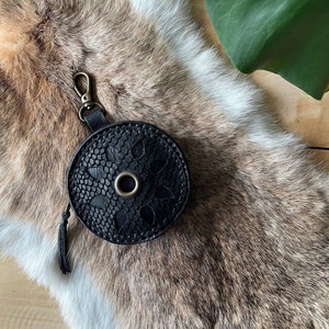 Op de afbeelding: Een zwarte, ronde leren tas met een bloemmotief en een metalen ring. De tas heeft een kleine kwast en een metalen sluiting. De tas ligt op een bruin en wit harig oppervlak.