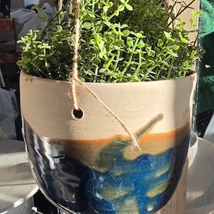 Könnte beinhalten: Ein hängender Keramik-Pflanztopf mit zweifarbigem Design. Die obere Hälfte ist cremefarben, während die untere Hälfte ein glänzendes, abstraktes Muster in Blau-, Grün- und Brauntönen aufweist. Der Topf ist mit leuchtend grünem Laub gefüllt und an einer natürlichen Schnur aufgehängt.